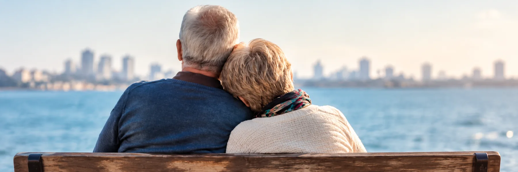 Pareja de pensionistas abrazada, de espaldas, mirando el mar y la ciudad desde un banco